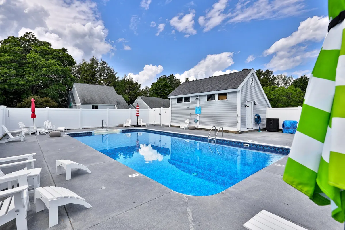 An inviting outdoor swimming pool surrounded by lounge chairs, vibrant poolside umbrellas, a changing room building, and lush trees. The setting is relaxed and cheerful, suggesting a communal space perfect for unwinding after a day of activity.