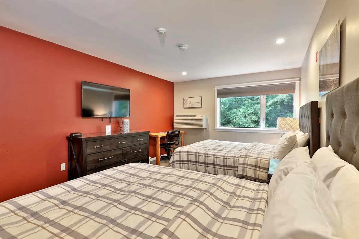 This photo captures the main studio area with two plush queen beds as the focal point, a wall-mounted television, a comfortable desk with chair positioned by a large window, and natural light streaming into the space. The vibe is cozy and welcoming, ideal for relaxation, working, or simply enjoying the view.