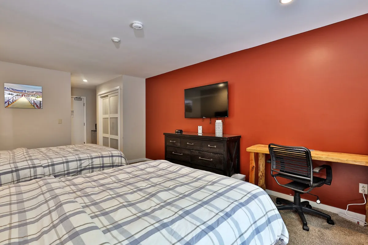The image features a main studio area with two beds, a TV mounted on the wall, a wooden desk with a chair, and a warm red accent wall. The space is inviting and set up for both relaxation and light productivity.