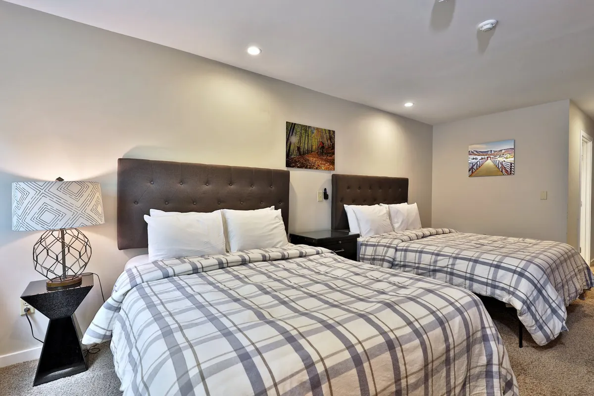 The image showcases a warm and inviting studio interior, centered around two queen beds with cozy plaid bedding. A soft decorative lamp casts a gentle glow over the beds, and tasteful wall art adds a touch of Vermont charm. The space feels intimate and comfortable, ideal for relaxation or unwinding after a day on the slopes.